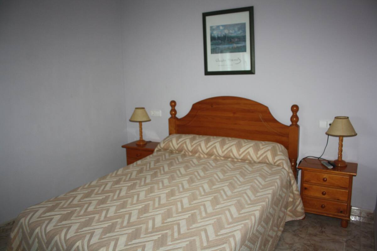 A bedroom with a wooden bed, two bedside tables with lamps, a framed picture on the wall, and a patterned bedspread.