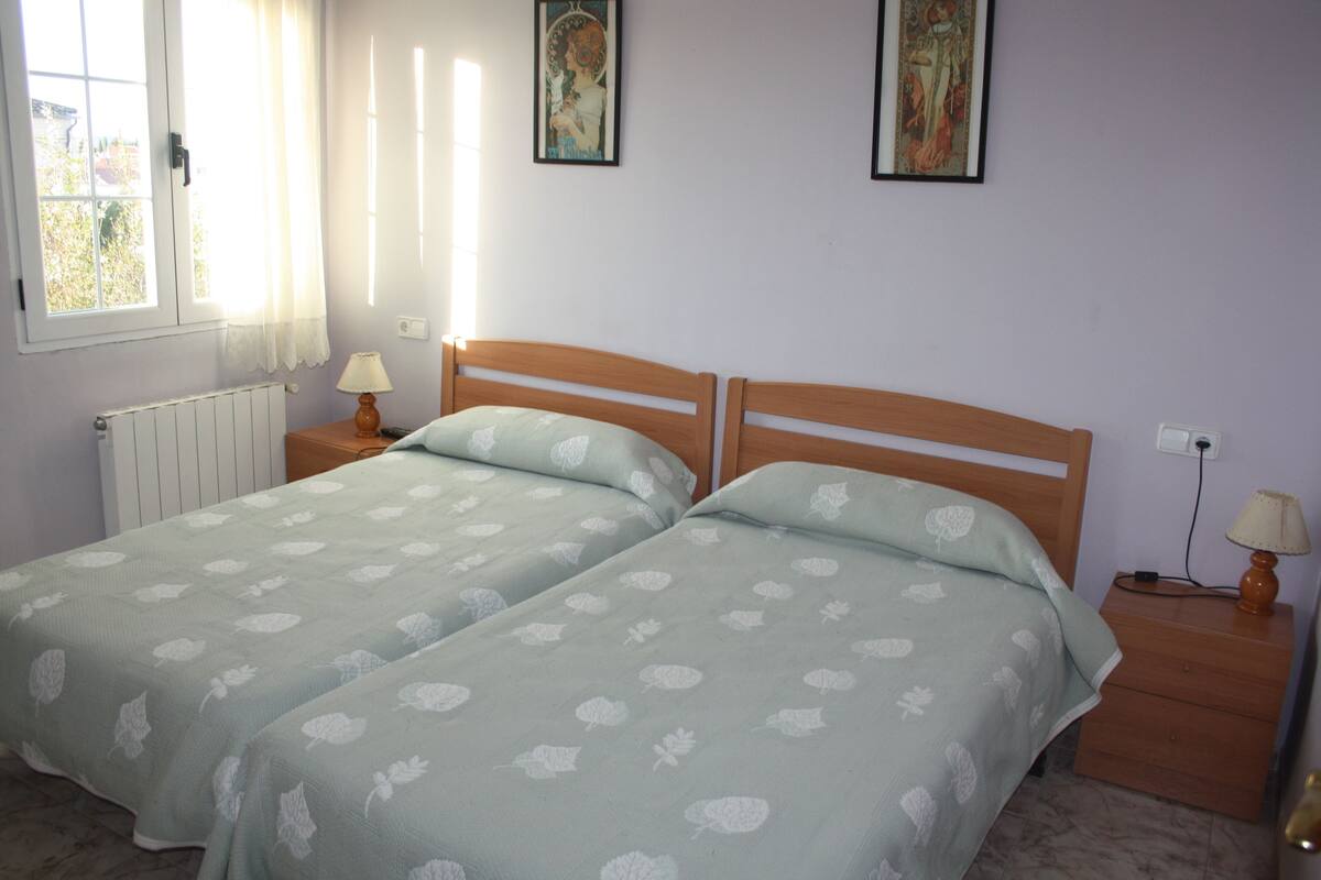 A bedroom with two single beds, a window with curtains, a radiator, and a nightstand with a lamp.