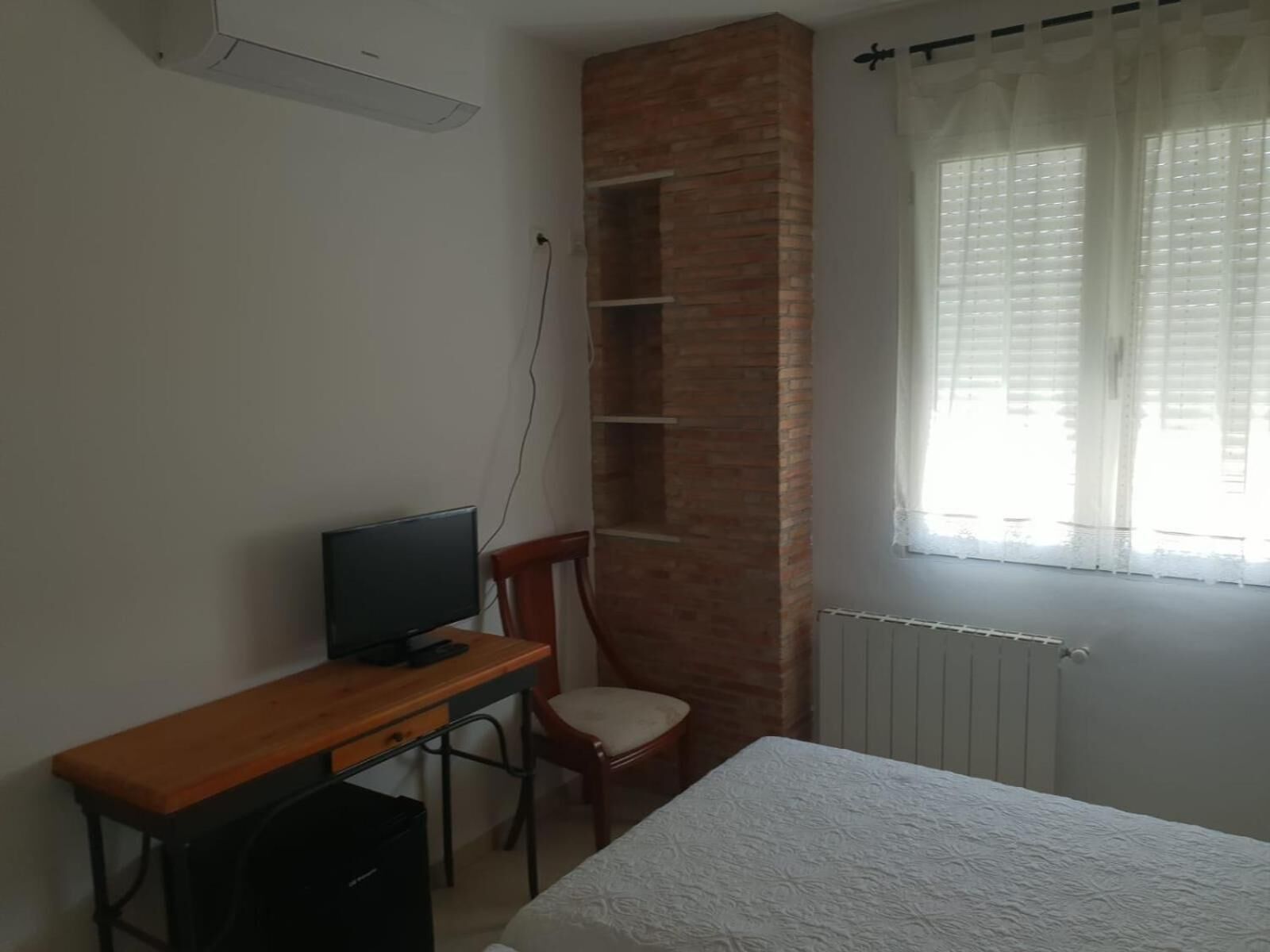 A bedroom with a bed, a desk with a computer, a chair, a brick accent wall, and a window with curtains.