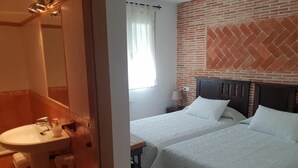 Una habitación de hotel con pared de ladrillo, una cama con ropa blanca, una mesita de noche con lámpara y un baño con lavabo y espejo.