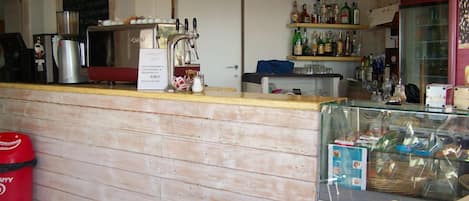 A bar area with a wooden counter, shelves with bottles, a refrigerator, and a trash bin.