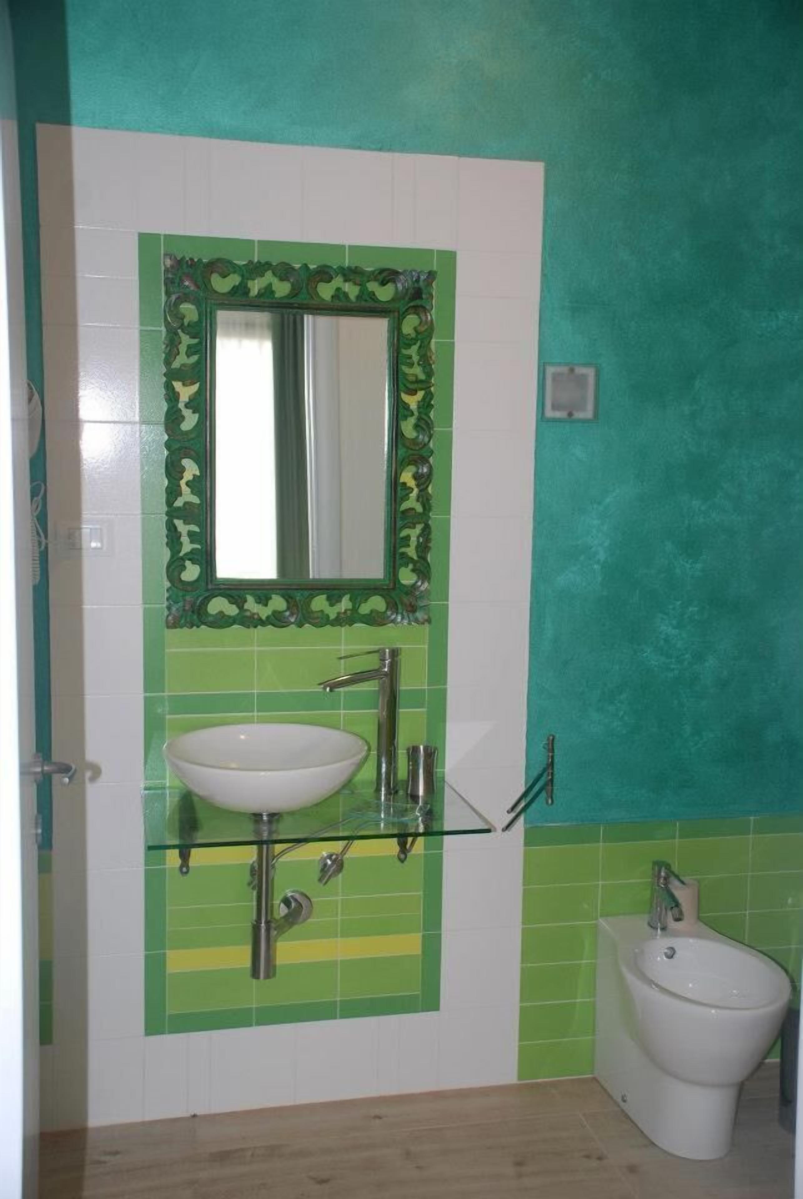 A bathroom with a green and white tiled wall, a white sink with a glass enclosure, a mirror with a decorative frame, and a white bidet.