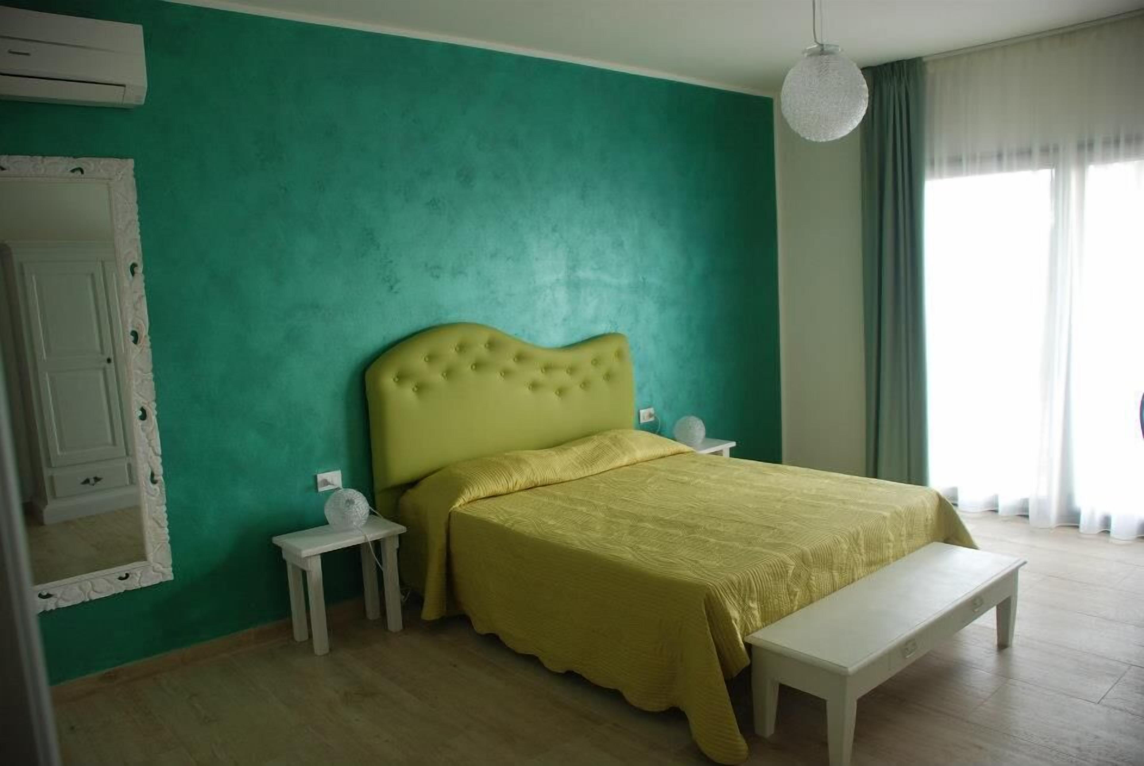 A bedroom with a green wall, a yellow bedspread, a white bench, and a white bedside table with a lamp.