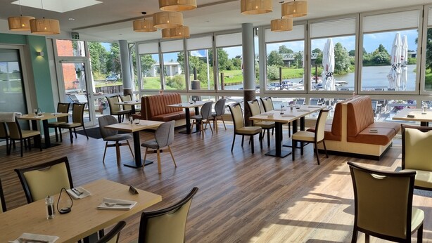 A spacious restaurant with large windows offering views of a lake and bridge, wooden tables and chairs, and hanging bamboo lamps. - Hotel Restaurant Bootshaus (Achim)