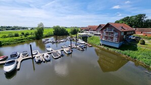 A riverside marina with boats docked, a building with balconies, and a green landscape in the background. - Hotel Restaurant Bootshaus (Achim)