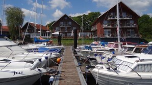 A marina with several boats docked, a wooden walkway, and a building with multiple balconies in the background. - Hotel Restaurant Bootshaus (Achim)