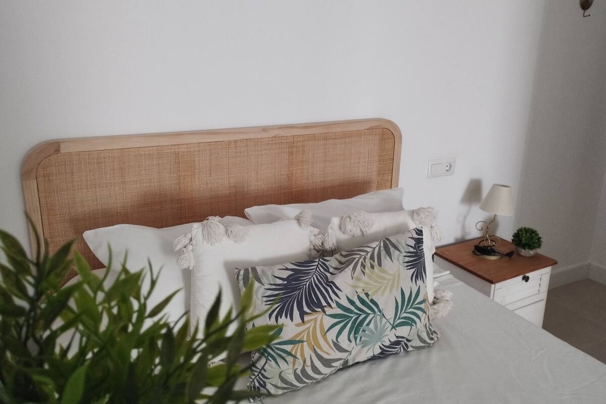 Una cama con cabecera de madera, ropa de cama blanca y un cojín con estampado floral. Hay una mesita de noche con una lámpara y una planta pequeña junto a la cama.