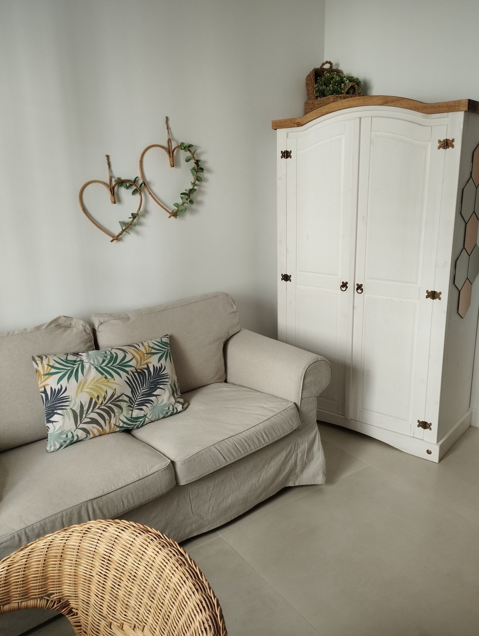 A neutral-toned living room with a beige sofa, a wicker chair, and a white wardrobe with decorative handles.