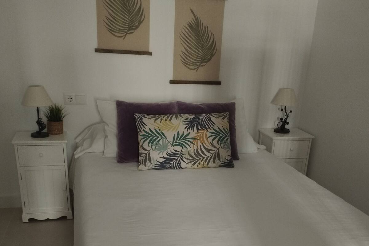 Una cama bien hecha con edredón blanco y almohada de estampado de hojas coloridas. Hay dos mesitas de noche, cada una con una lámpara y una pequeña planta.