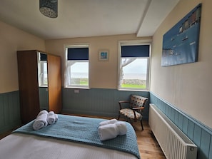 A bedroom with a bed, two windows, a chair, a radiator, and a painting on the wall.
