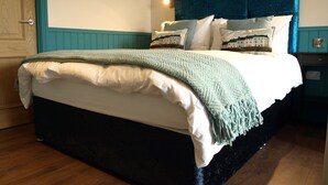 Una cama bien tendida con una manta verde y sábanas blancas, un cabecero oscuro y un cuadro en la pared.