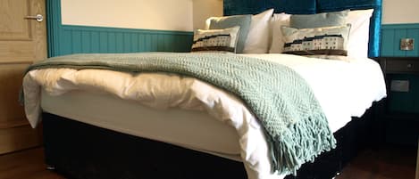 Una cama bien tendida con una manta verde y sábanas blancas, un cabecero oscuro y un cuadro en la pared.