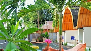 A pool area with lounge chairs, a small garden, and a building with a tiled roof. - Macoa Village (Gili Trawangan)