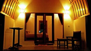 A cozy room with a bed, a small table, and a chair. There are two wall-mounted lights and a door leading to another room. - Macoa Village (Gili Trawangan)