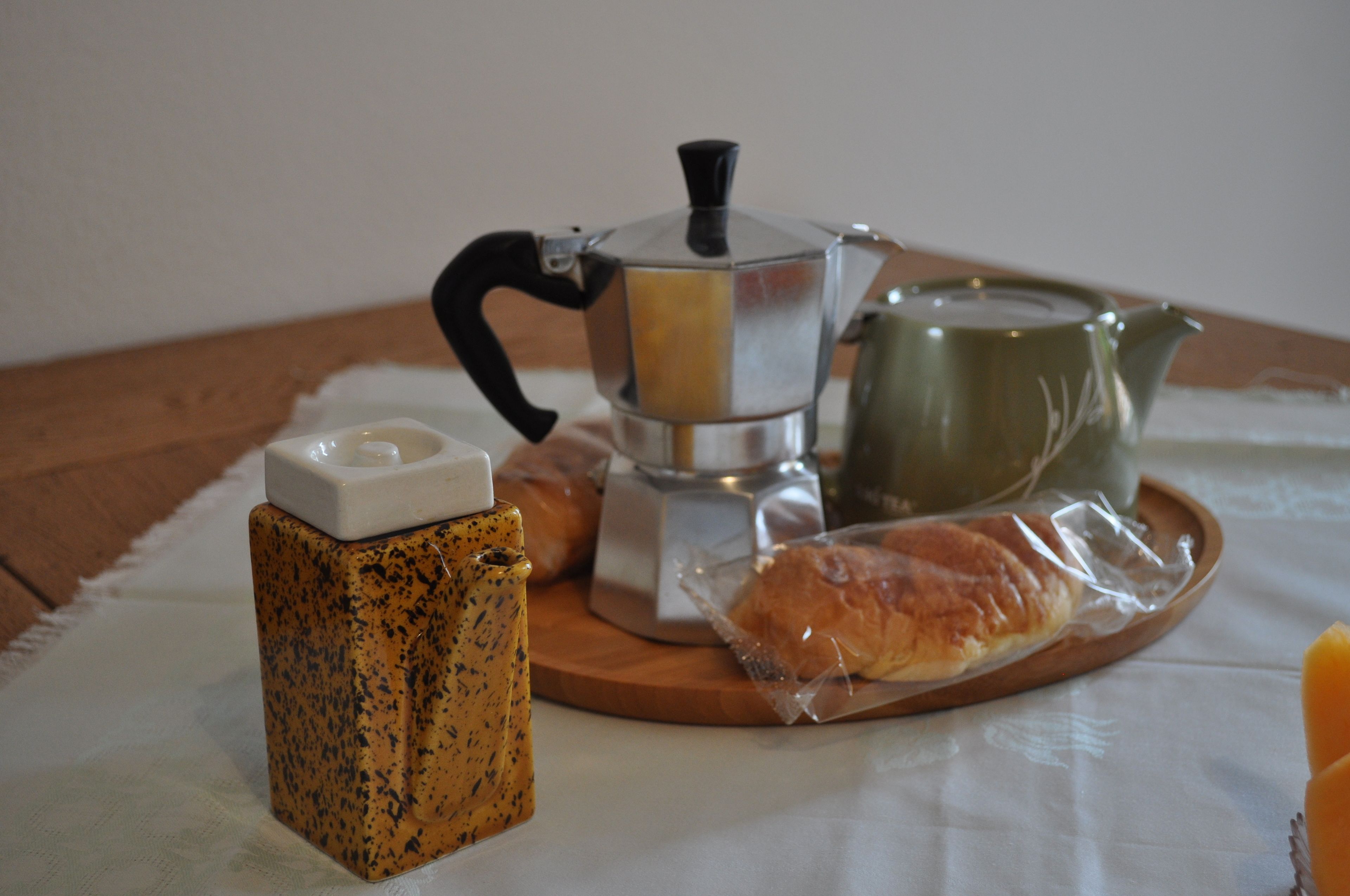 A coffee maker, a green teapot, and a croissant on a wooden tray.