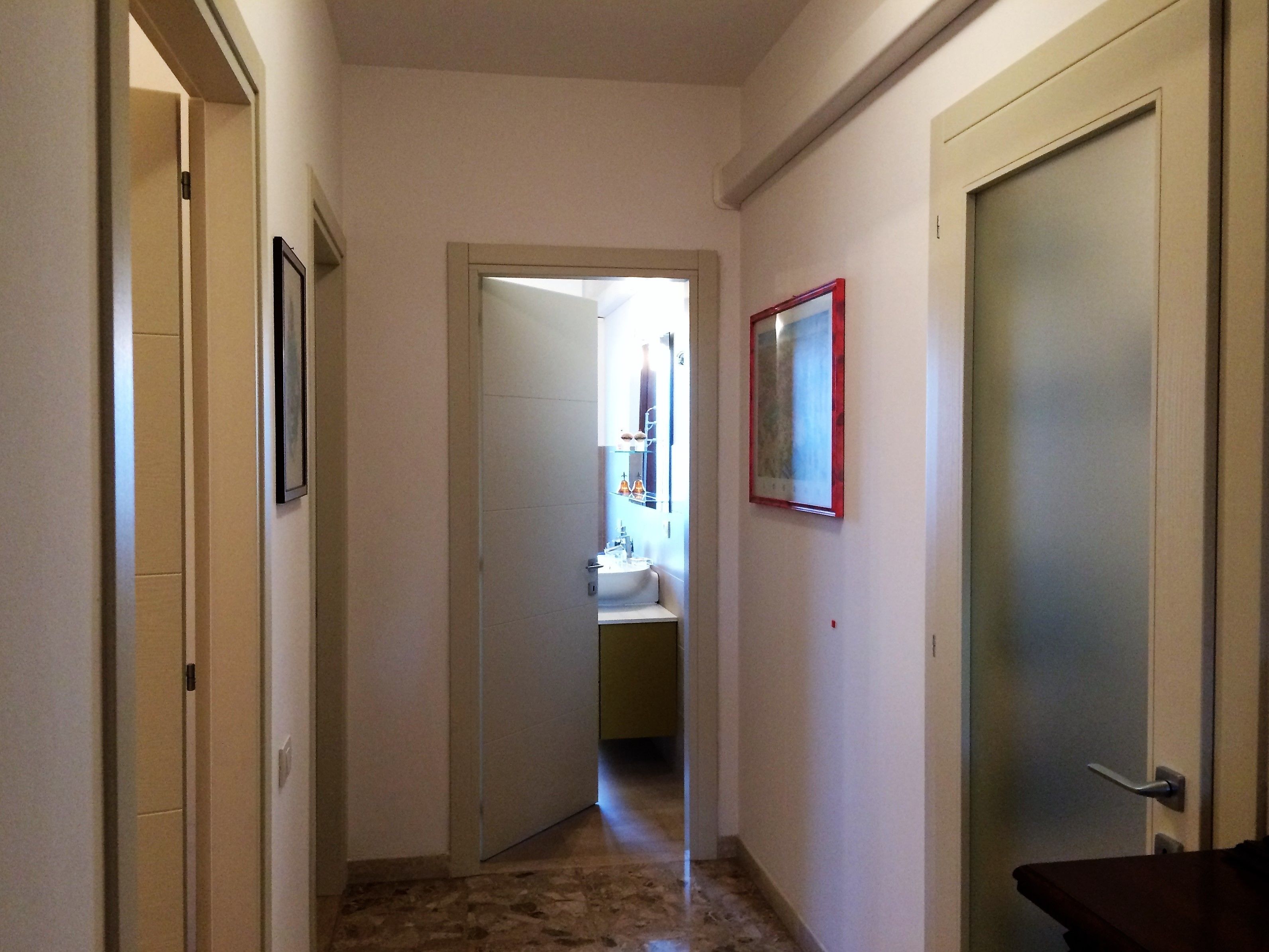 Ein schmaler Korridor mit einer Tür rechts, einem gerahmten Bild an der Wand und einem Blick in einen Raum am Ende.
