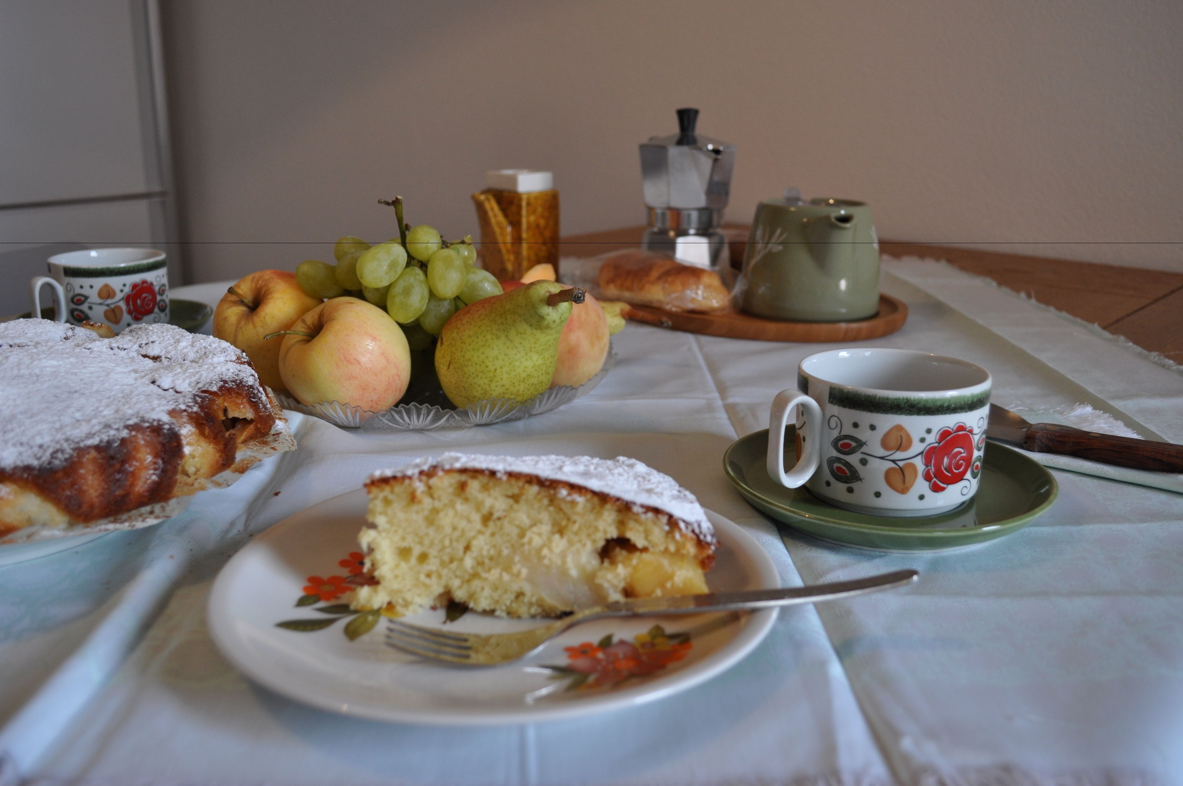 Ein Tisch mit Kuchenstück, Obstteller, Tasse und Teekanne.
