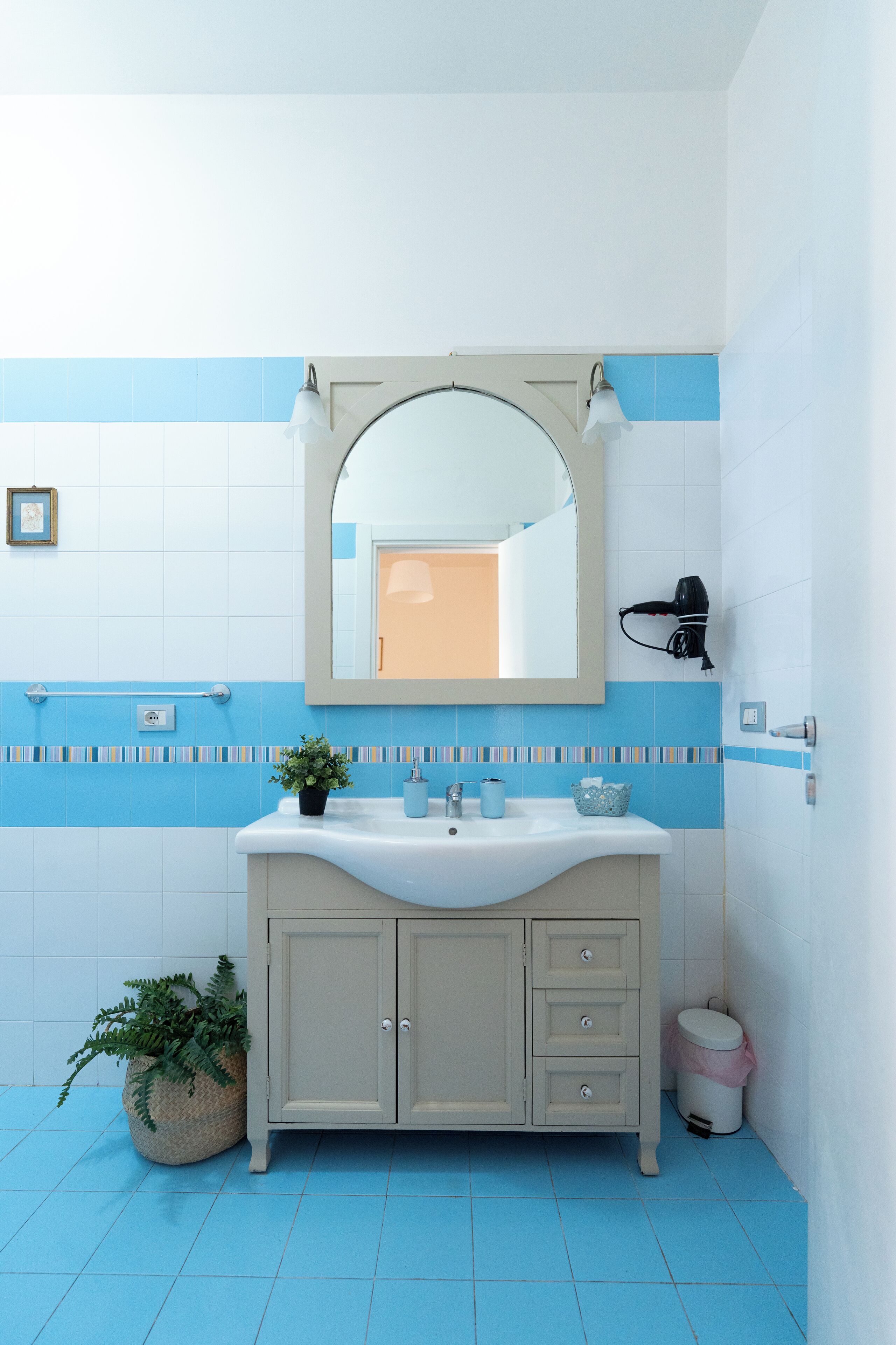A bathroom with a white sink, blue tiles, a mirror, a potted plant, and a trash bin.