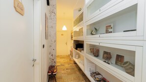 A narrow hallway with a tiled floor, a white door, and a shoe rack with red shoes. Shelves on the right display various items including books and decorative objects. - L'anfiteatro di Pompei B&B (Pompei)