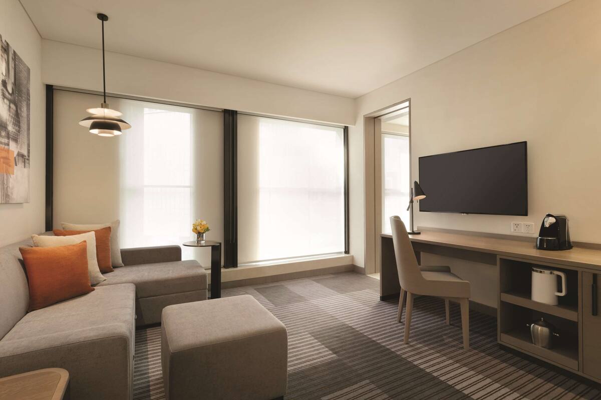 A modern hotel room with a sofa, ottoman, desk, chair, television, and a coffee maker.