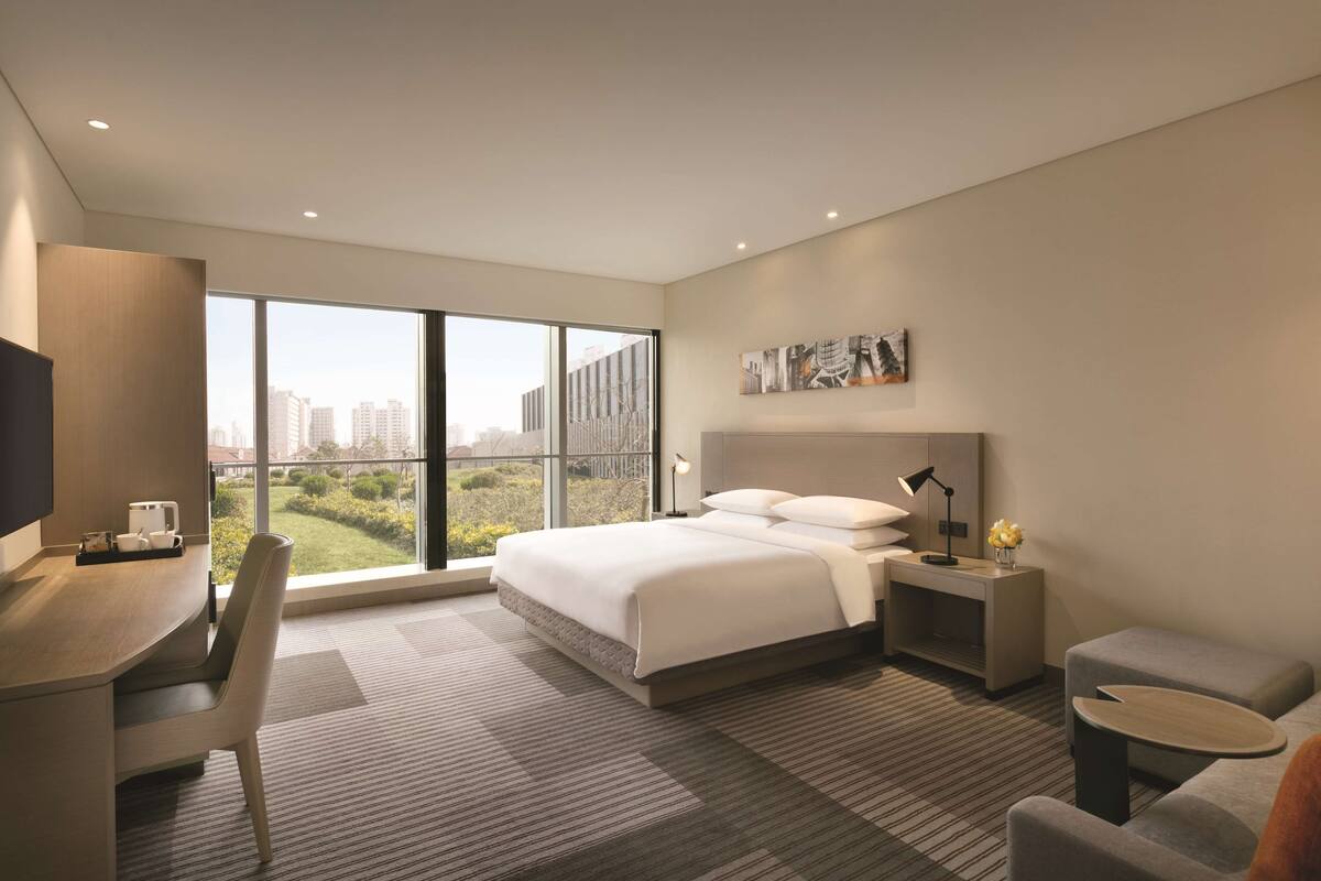 A modern hotel room with a large bed, a desk, a chair, a sofa, and a view of the cityscape.