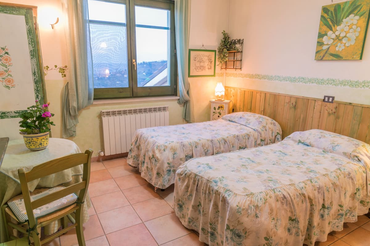 A room with two beds, a table, a chair, a window, a radiator, and a wall with a plant and a picture.