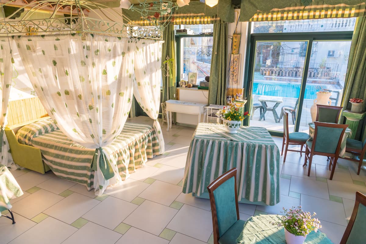 A room with a bed, a dining table, chairs, a small table, a potted plant, and a view of a pool.