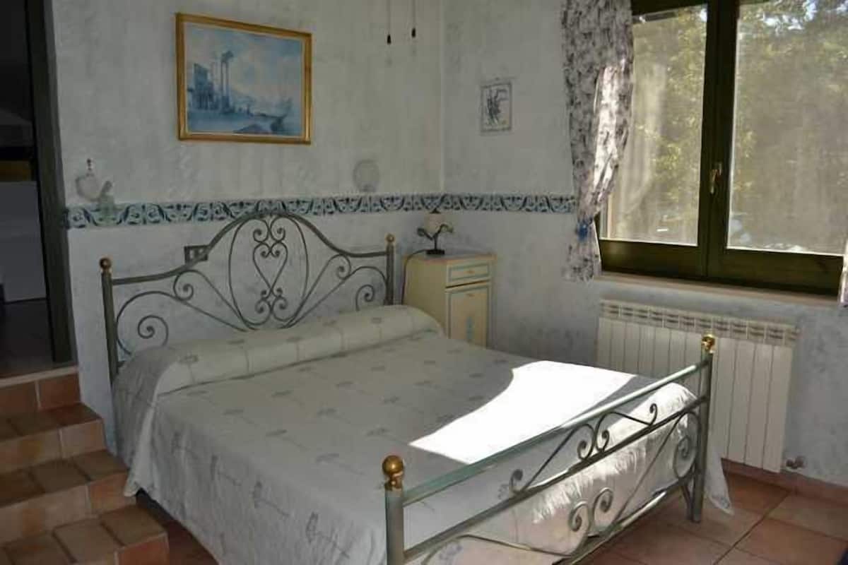A bedroom with a bed, a nightstand, a window with curtains, and a radiator.