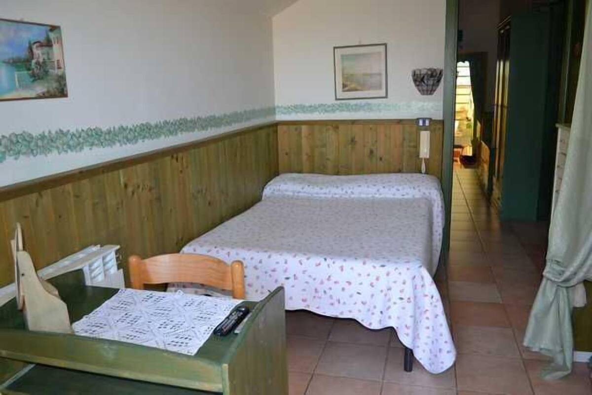 A small, single-bed room with a wooden headboard, a wooden chair, a table with a menu, and a painting on the wall.