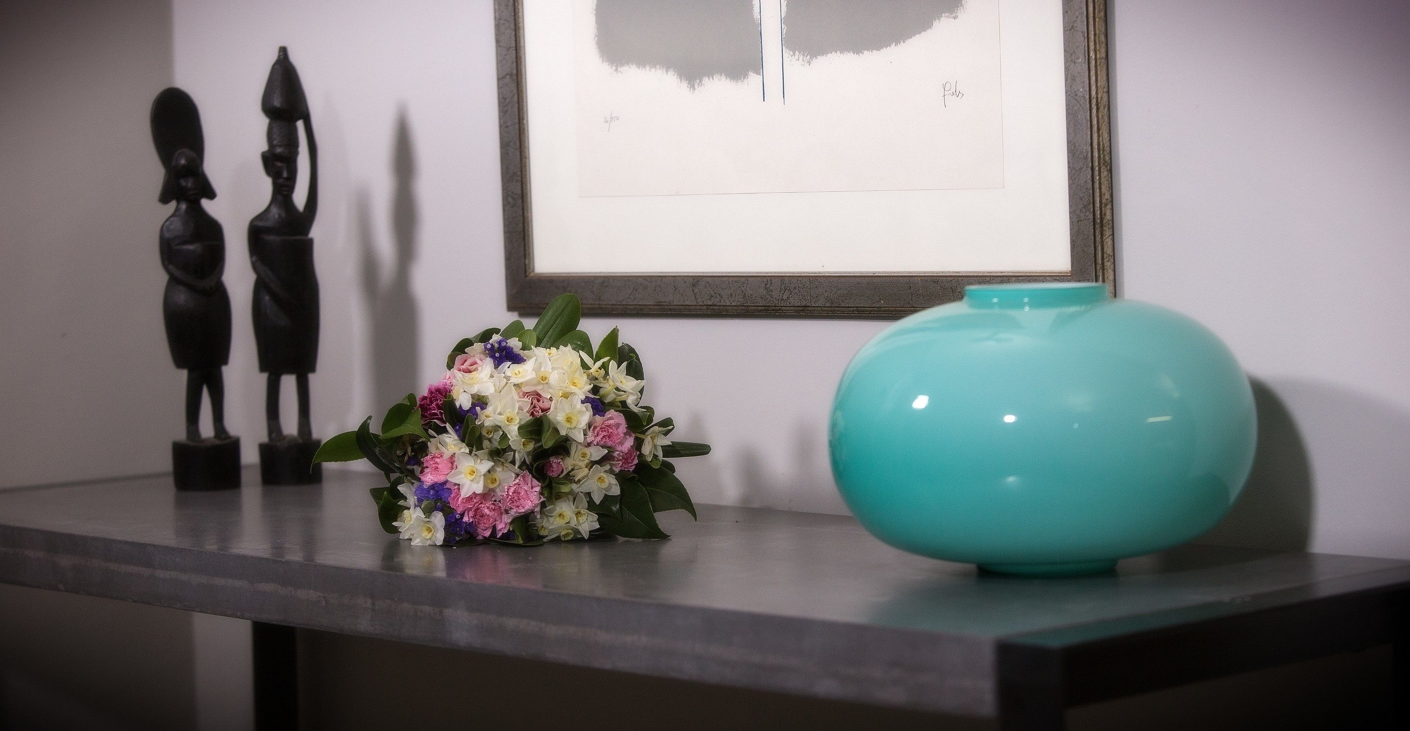 Una mesa con un jarrón de flores, un jarrón color turquesa y dos esculturas negras.