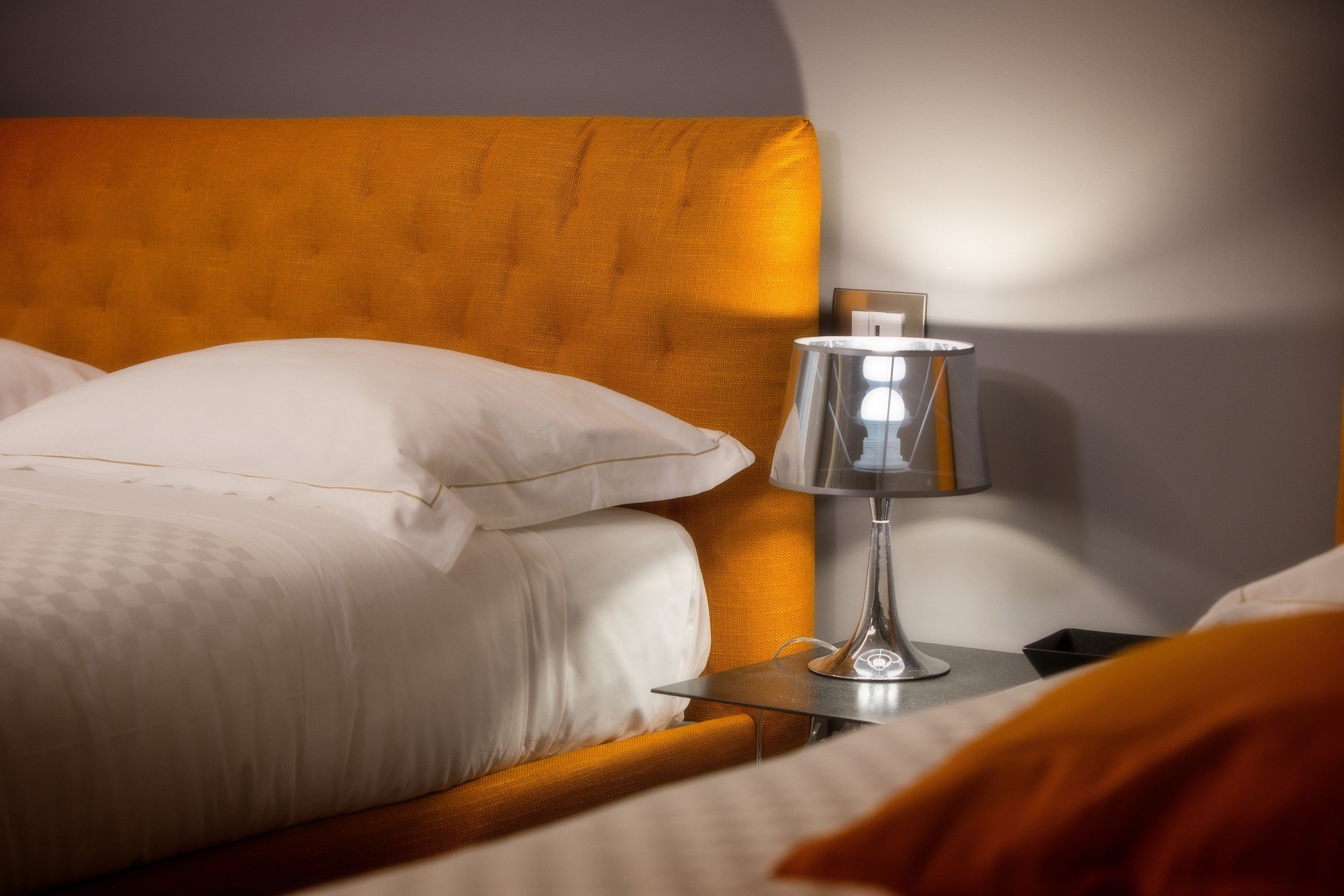 Una cama bien hecha con sábanas blancas y una única almohada blanca, una mesita de noche con una lámpara y un cabecero de madera.
