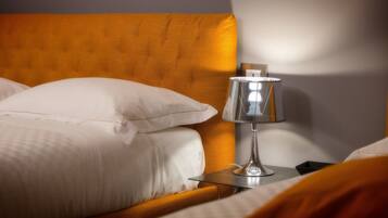 Una cama bien hecha con sábanas blancas y una única almohada blanca, una mesita de noche con una lámpara y un cabecero de madera.