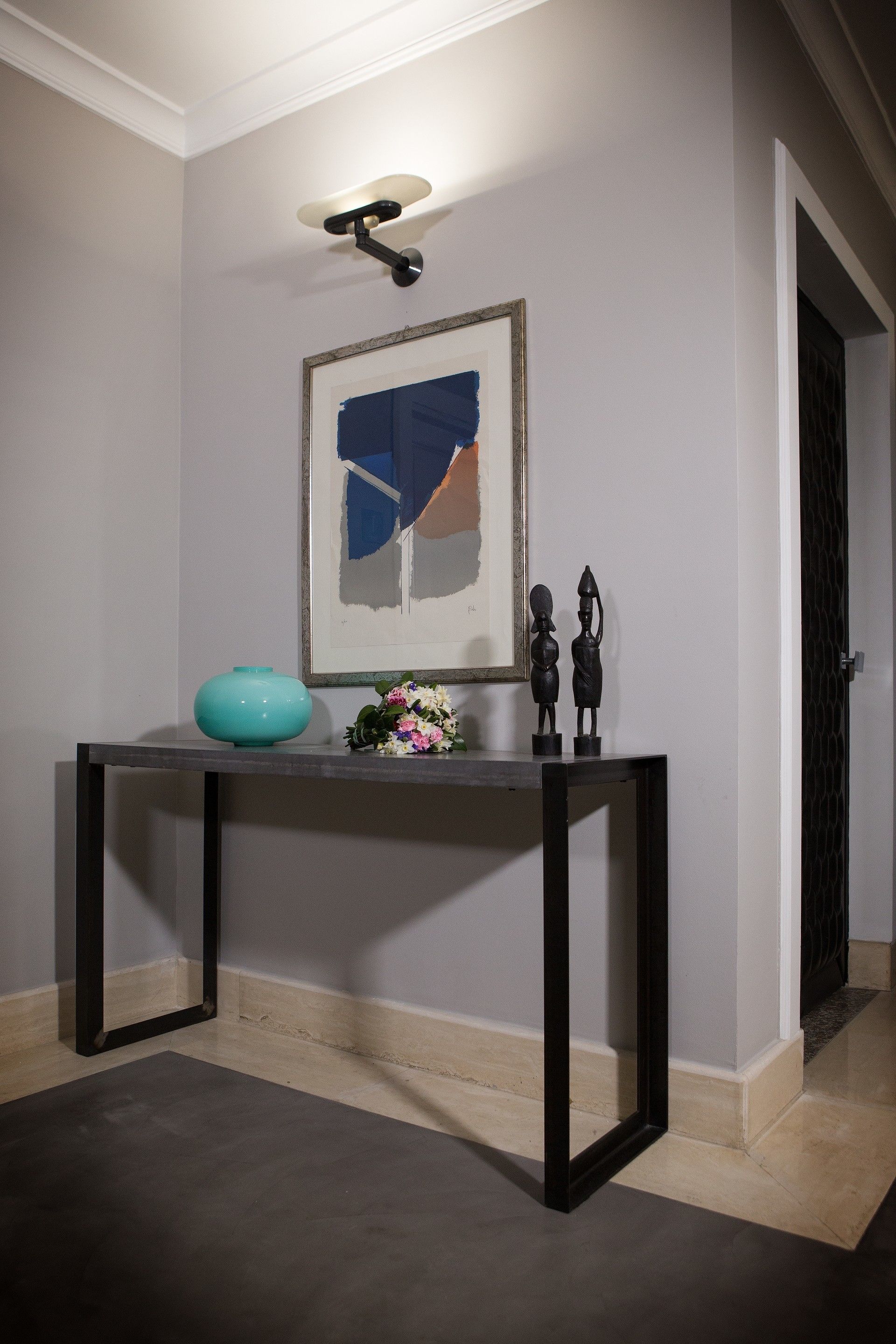 A hallway with a dark wooden console table, a framed abstract painting, a teal vase, and two black sculptures.