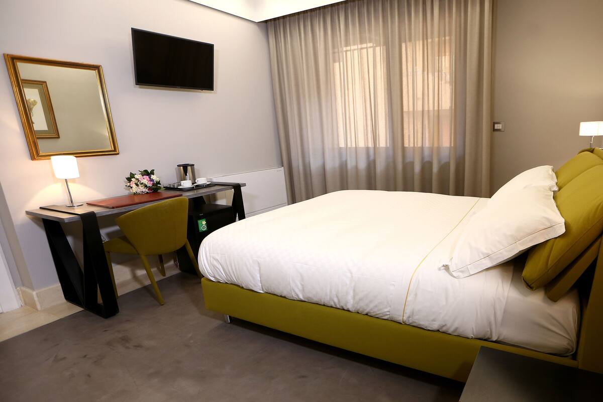 A hotel room with a bed, a desk, a chair, a TV, a lamp, and a window with curtains.