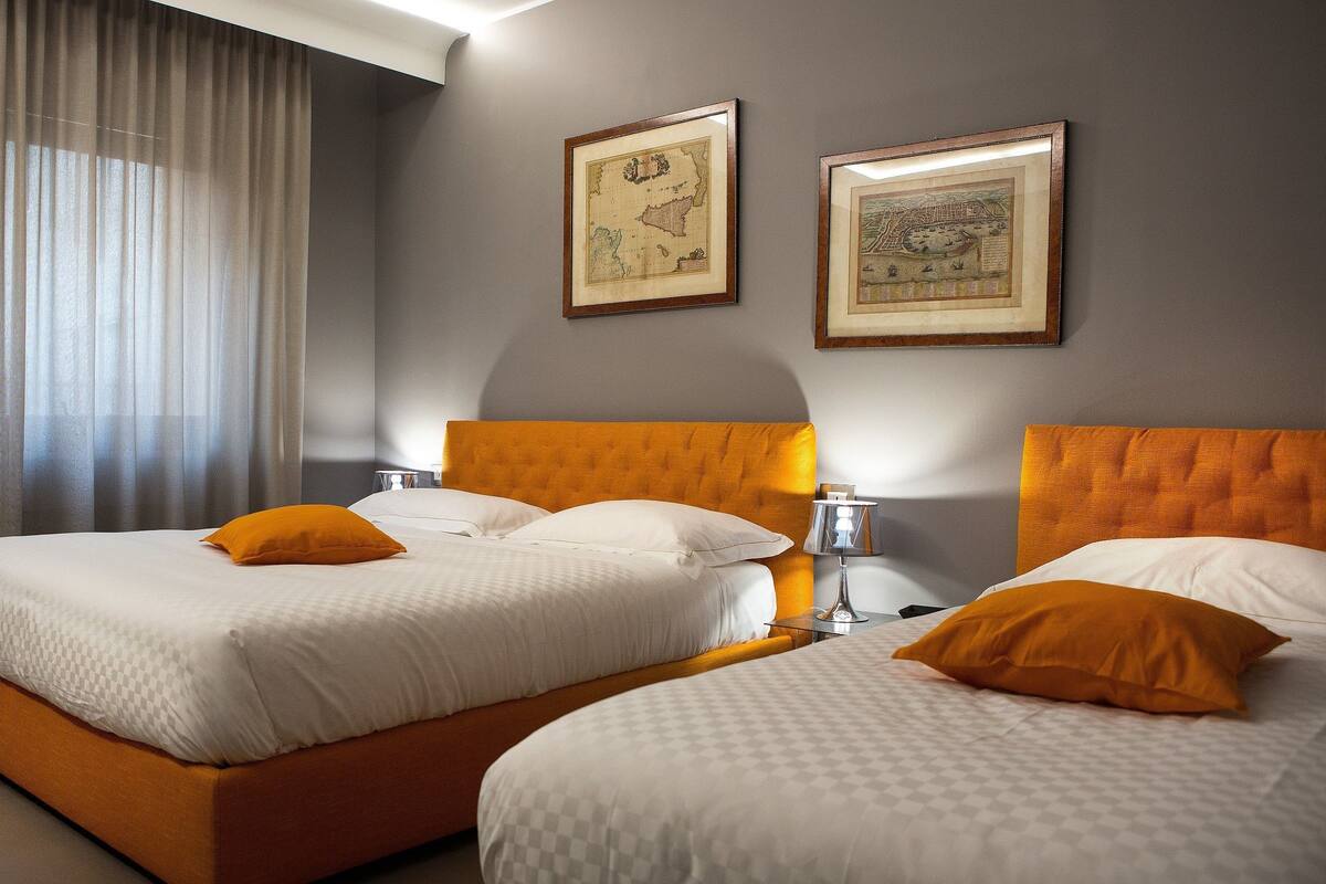 A hotel room with two beds, a large window with curtains, and two framed maps on the wall.