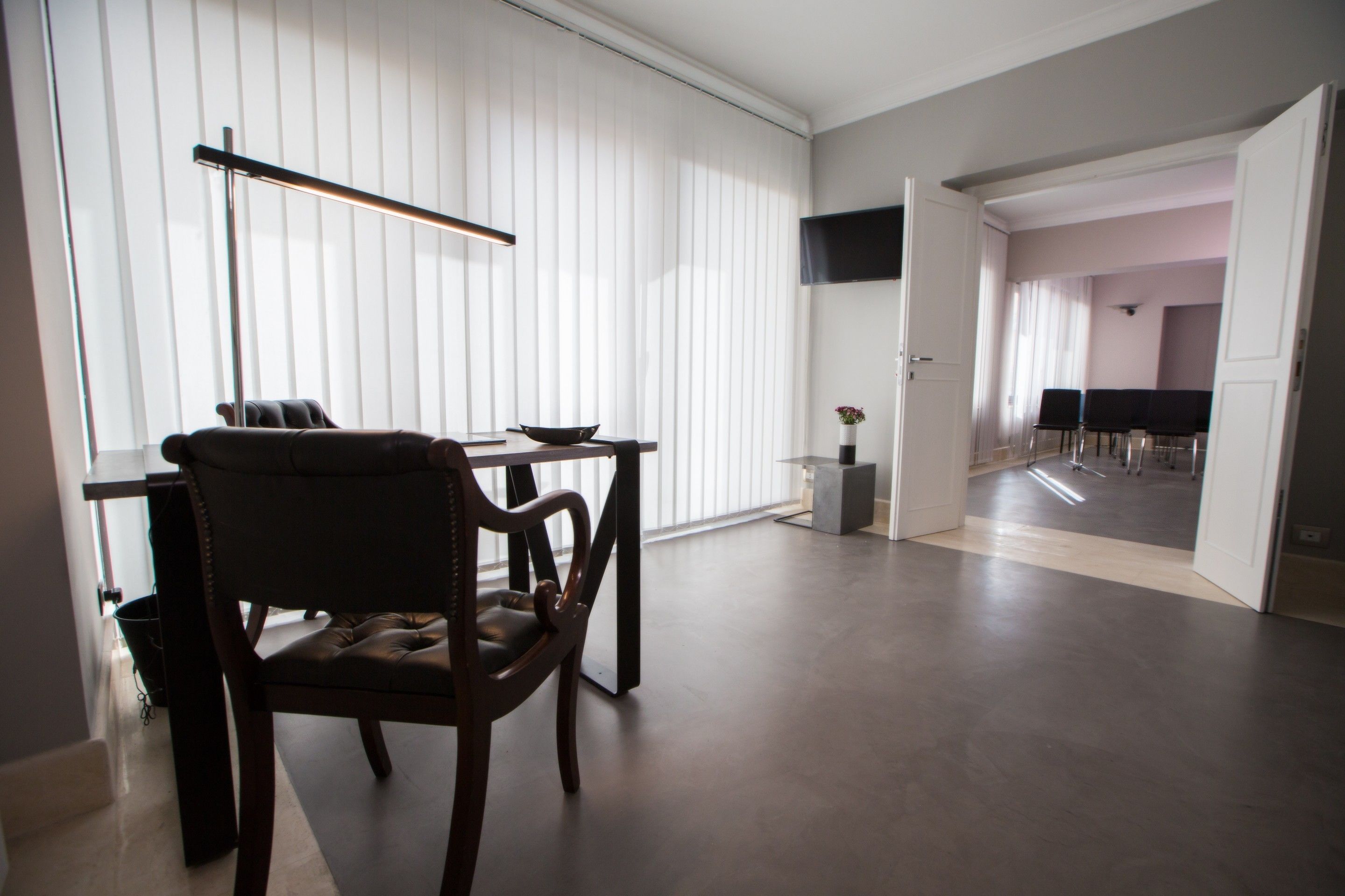 A modern living room with a dark wooden chair, a small table, and a floor lamp. There is a door leading to another room with a dining area.