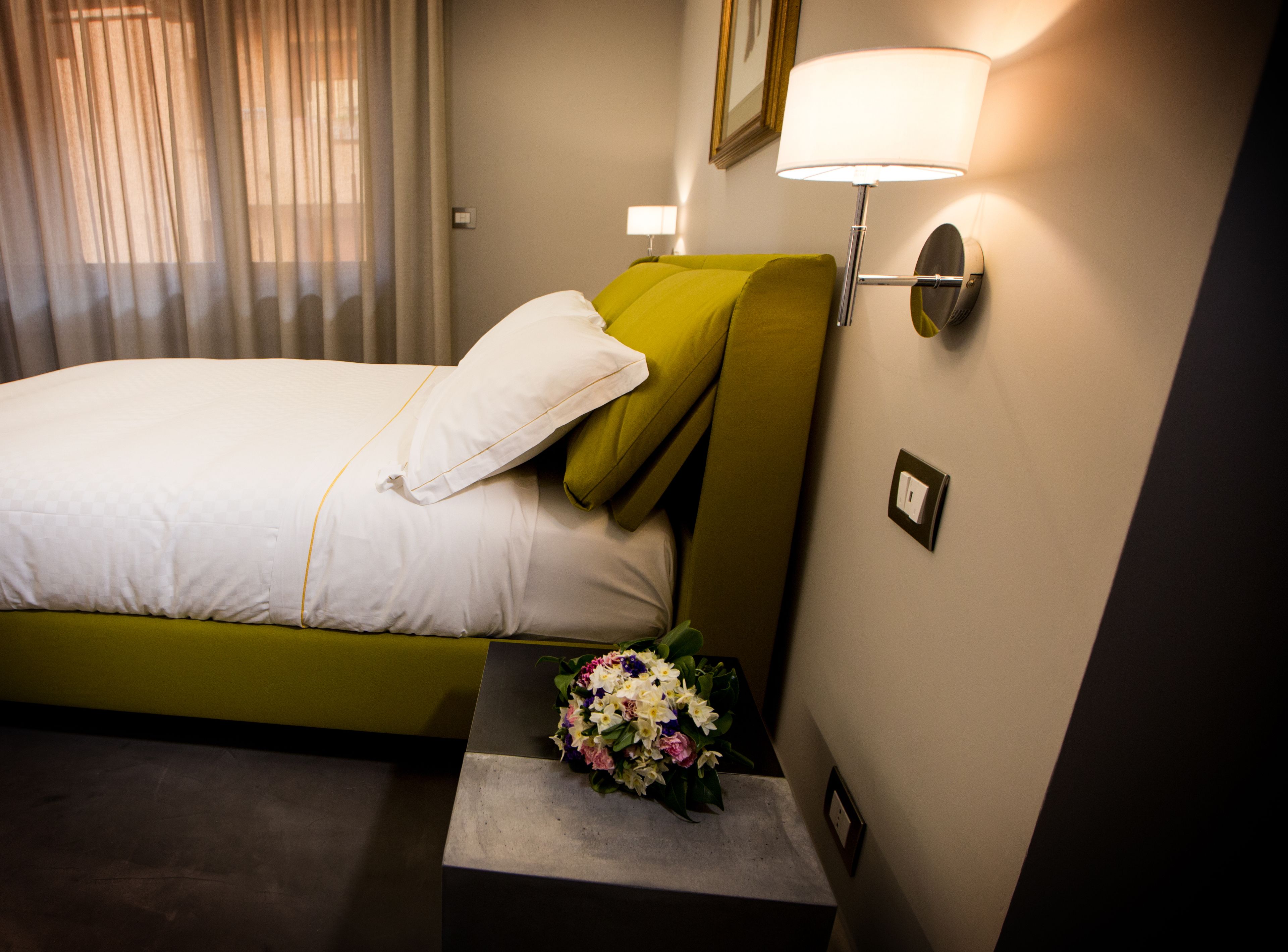 A hotel room with a bed, a nightstand with a flower arrangement, a lamp, and a wall-mounted light fixture.