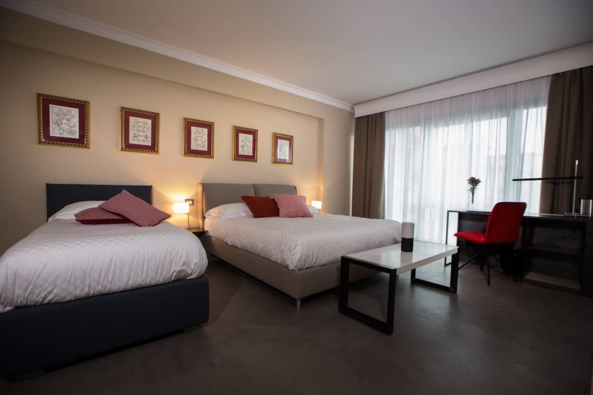 A hotel room with two beds, a desk, and a chair. There are framed pictures on the wall and a window with curtains.