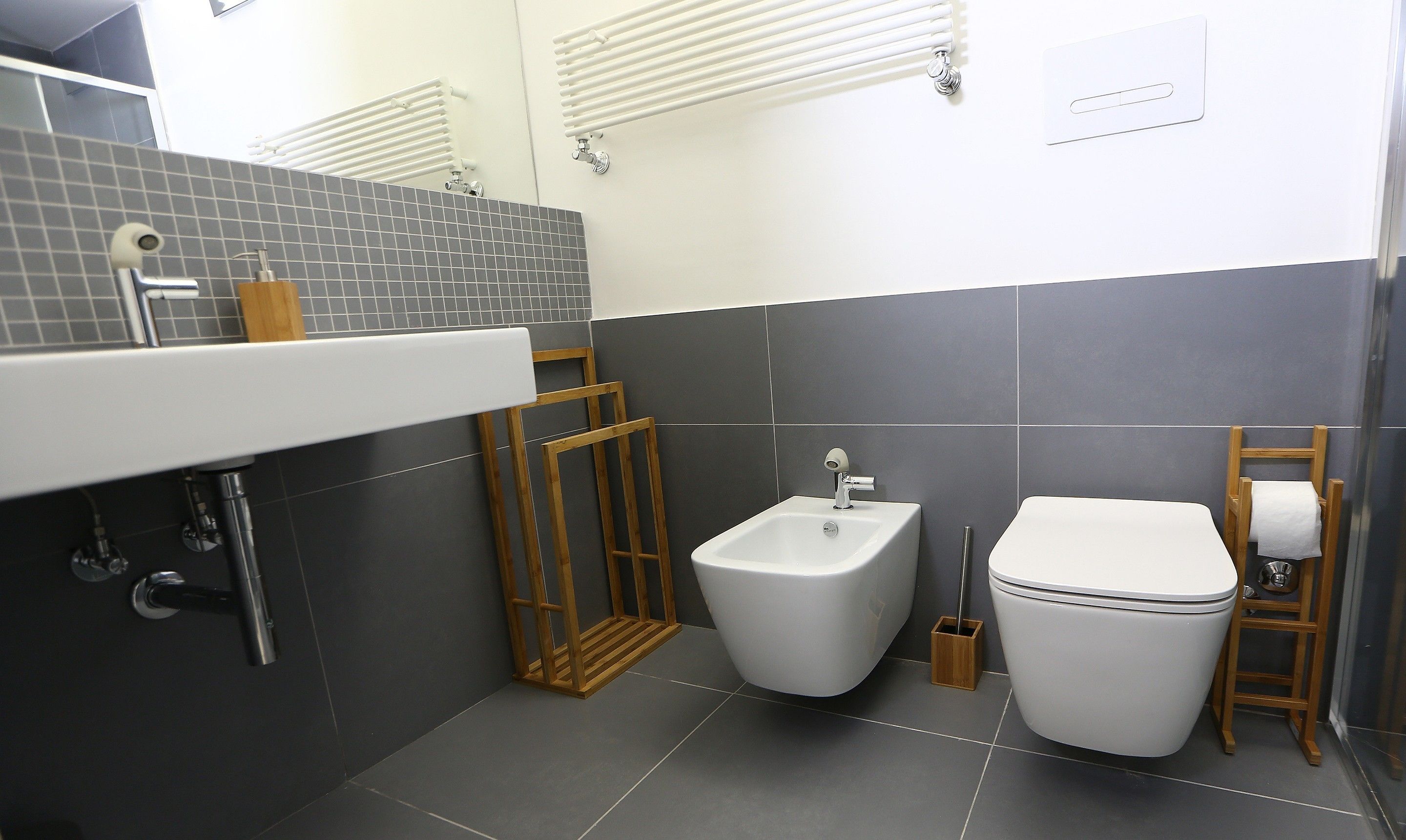 Un baño moderno con lavamanos, bidé y retrete blancos, baldosas grises y elementos de madera.