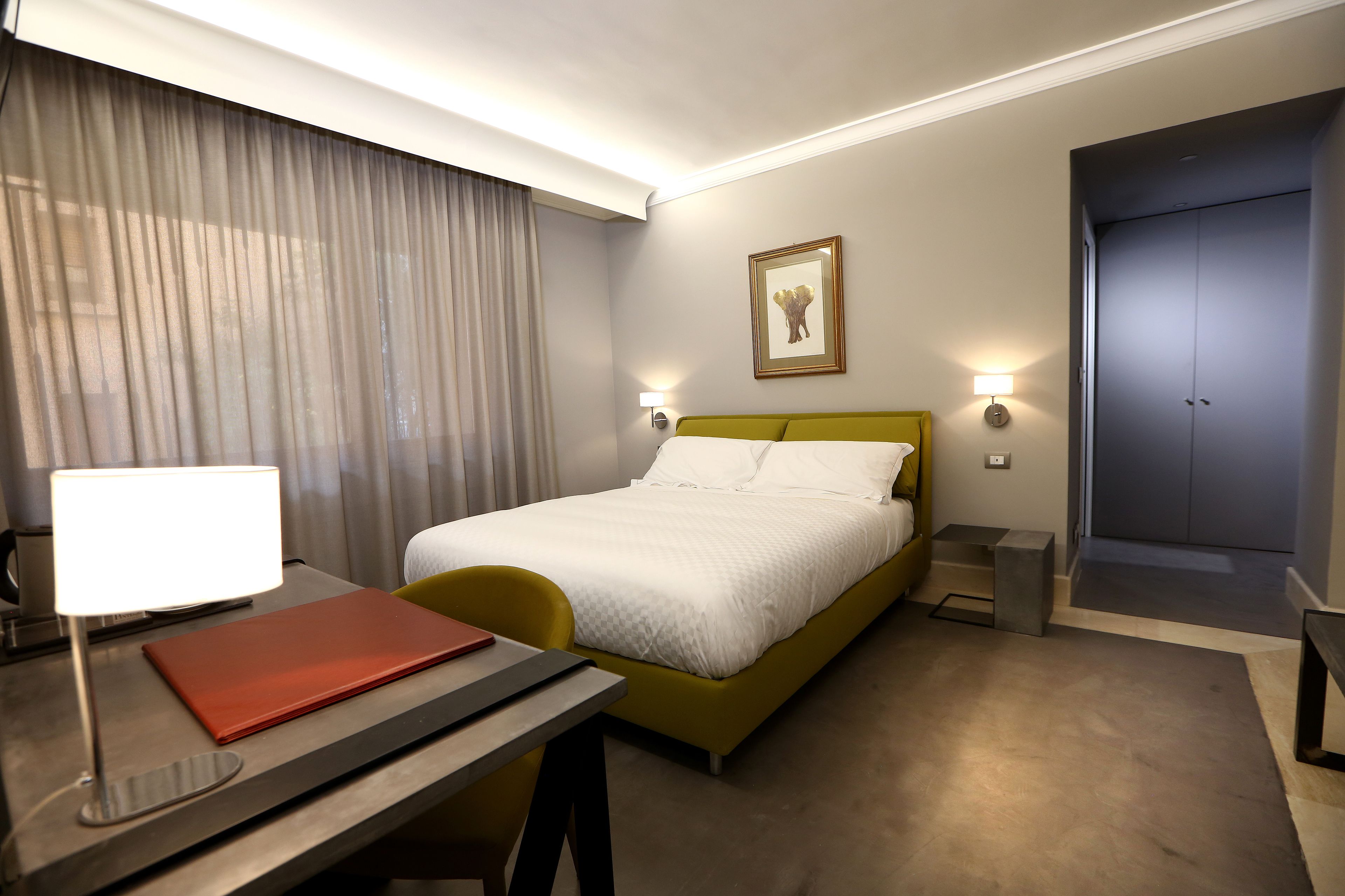 Habitación de hotel con cama, escritorio con lámpara, silla, mesita de noche y puerta.