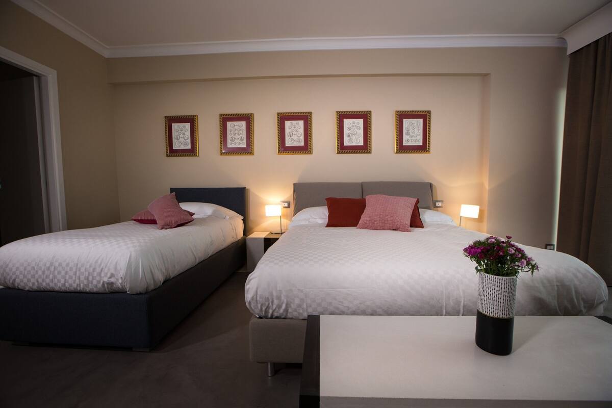 A hotel room with two beds, a bedside table, a nightstand, and a wall with framed pictures.