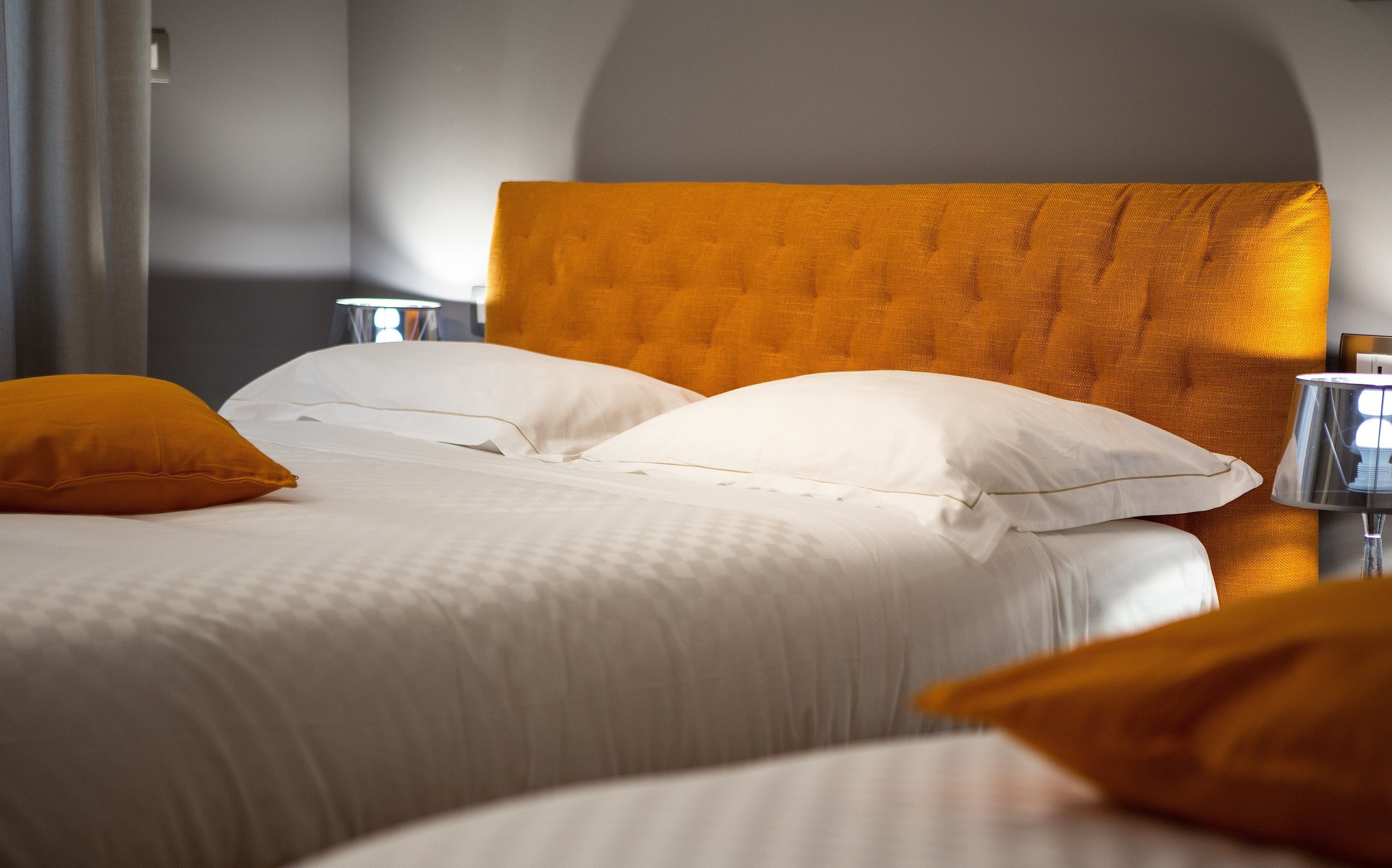 Ein ordentlich bezogenes Bett mit weißen Leinen und zwei orangefarbenen Kissen, ein Kopfteil in warmer Farbe und ein Nachttisch mit einer Lampe.