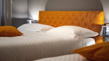 Ein ordentlich bezogenes Bett mit weißen Leinen und zwei orangefarbenen Kissen, ein Kopfteil in warmer Farbe und ein Nachttisch mit einer Lampe.