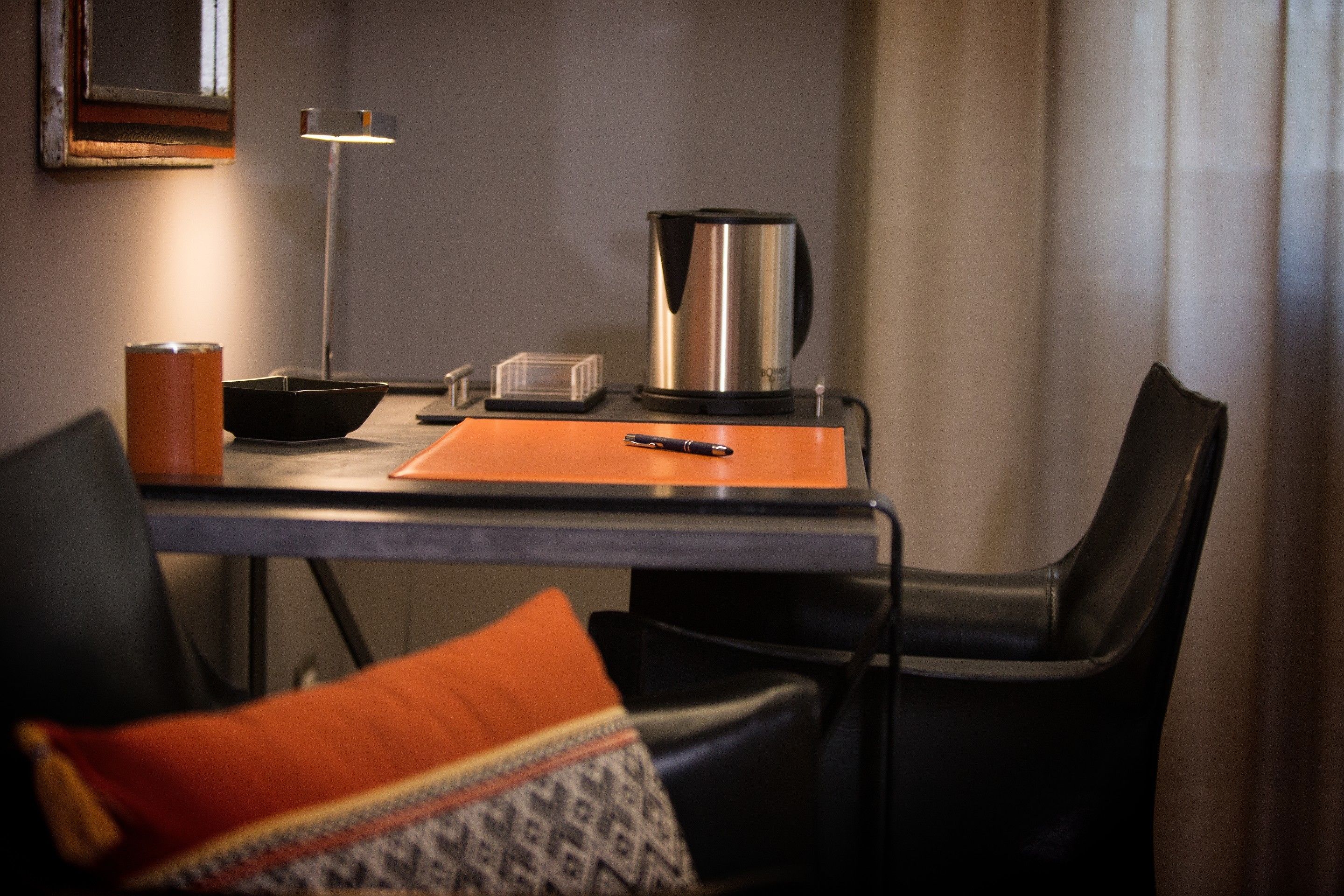 A small, modern room with a table, chairs, a kettle, a lamp, and a bed with an orange pillow.