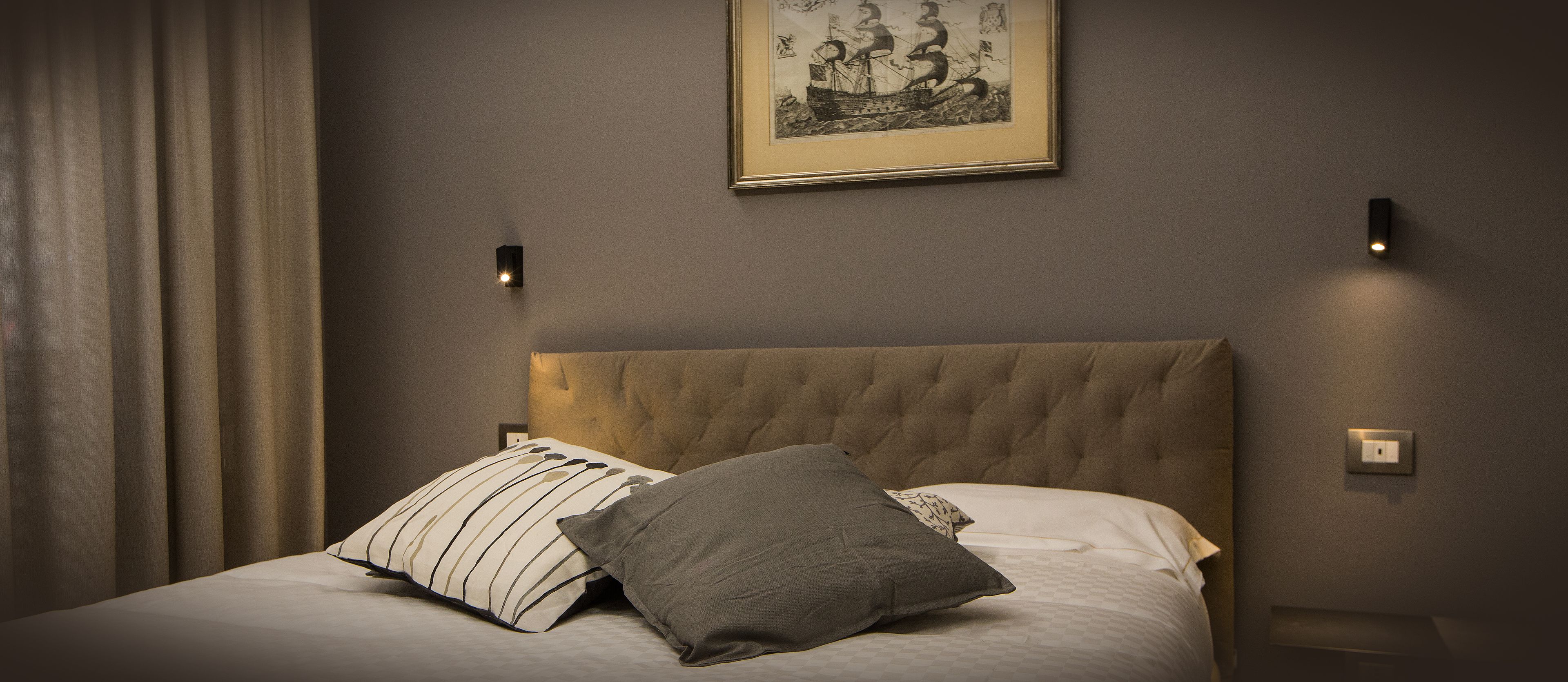 Un lit bien fait, avec un oreiller à rayures et un oreiller gris, une œuvre d’art encadrée au-dessus du lit, et deux lampes fixées au mur.
