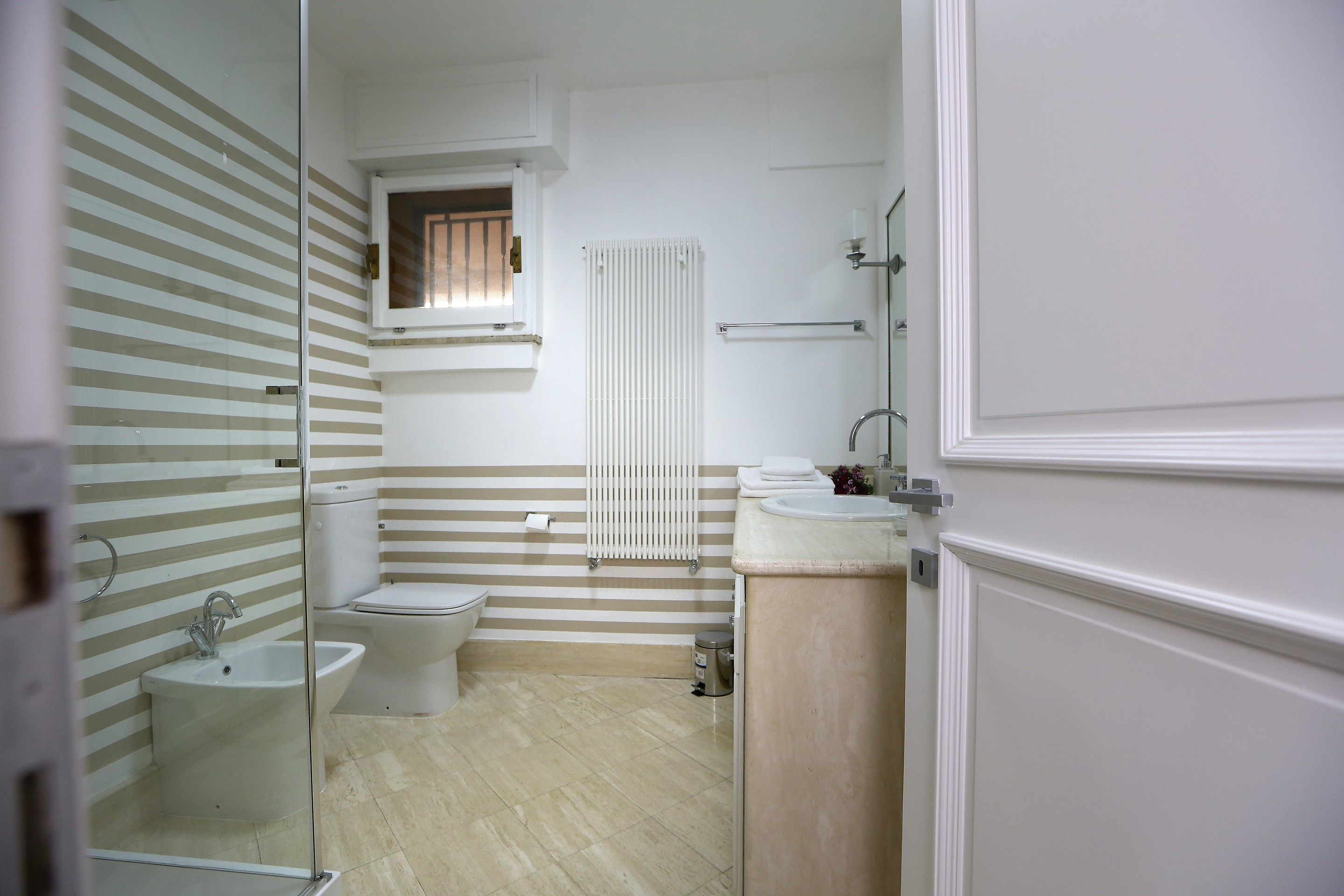 A bathroom with a toilet, bidet, and sink. There is a window with blinds, a towel rack, and a radiator.