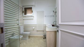 A bathroom with a toilet, bidet, and sink. There is a window with blinds, a towel rack, and a radiator.