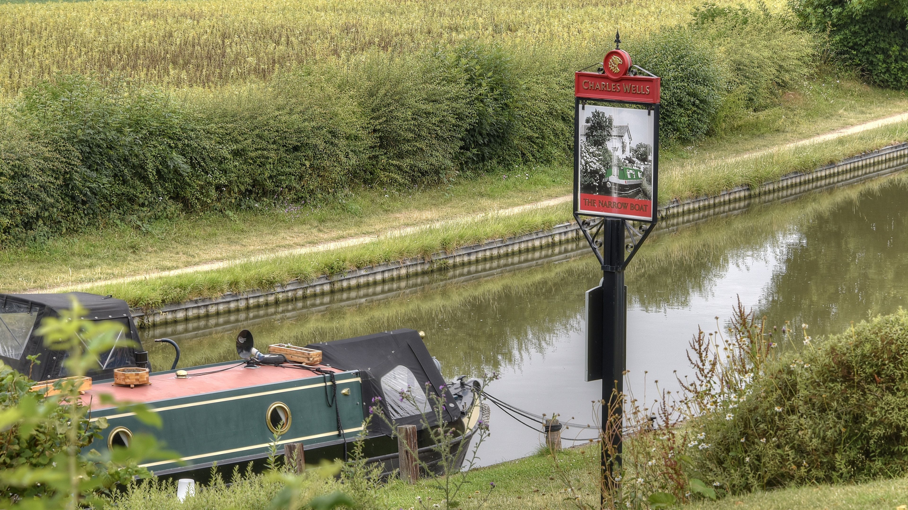 A narrow boat docked by a canal with a sign indicating 'Charles Wells' and 'The Narrow Boat'.