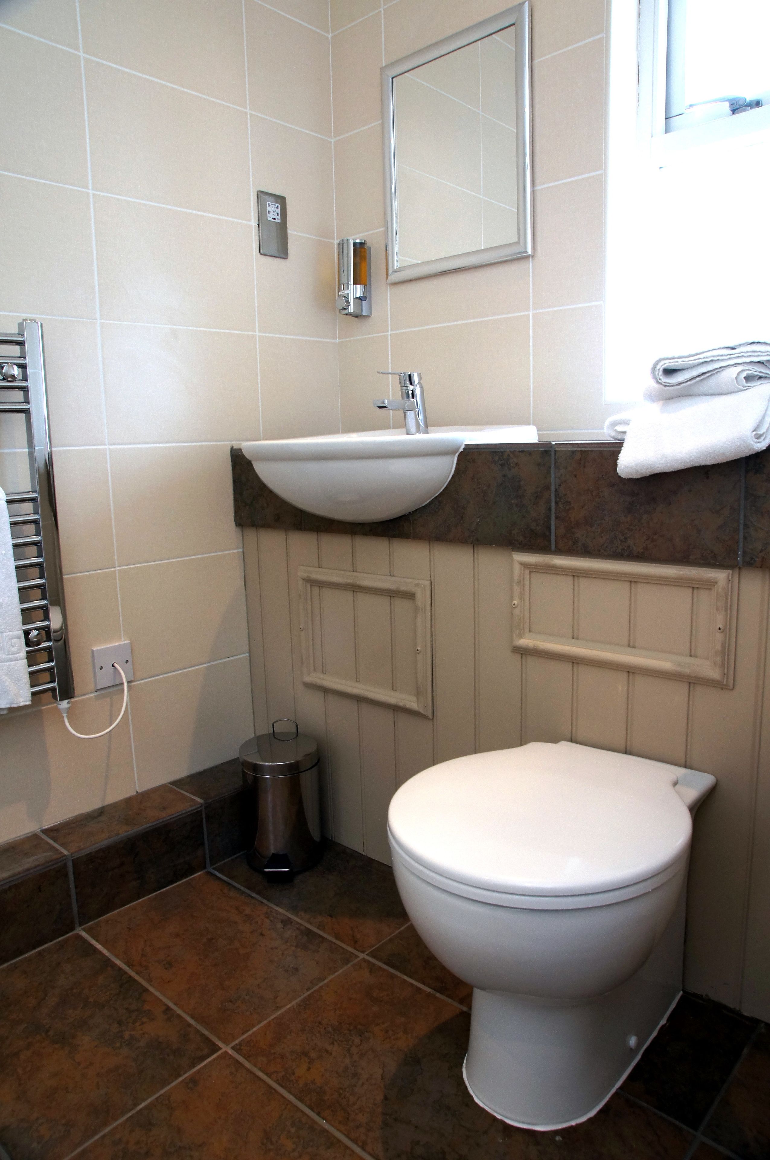 Et badeværelse med et hvidt toilet, en vask og et spejl. Der er en håndklædestang og en skraldespand.