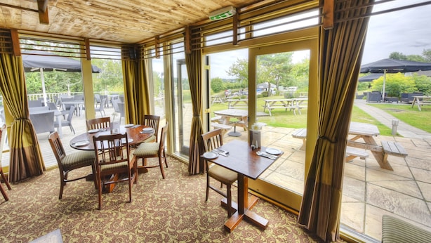 A dining area with wooden tables and chairs, large windows, and a view of the outdoor patio. - The Narrowboat At Weedon (Northampton)